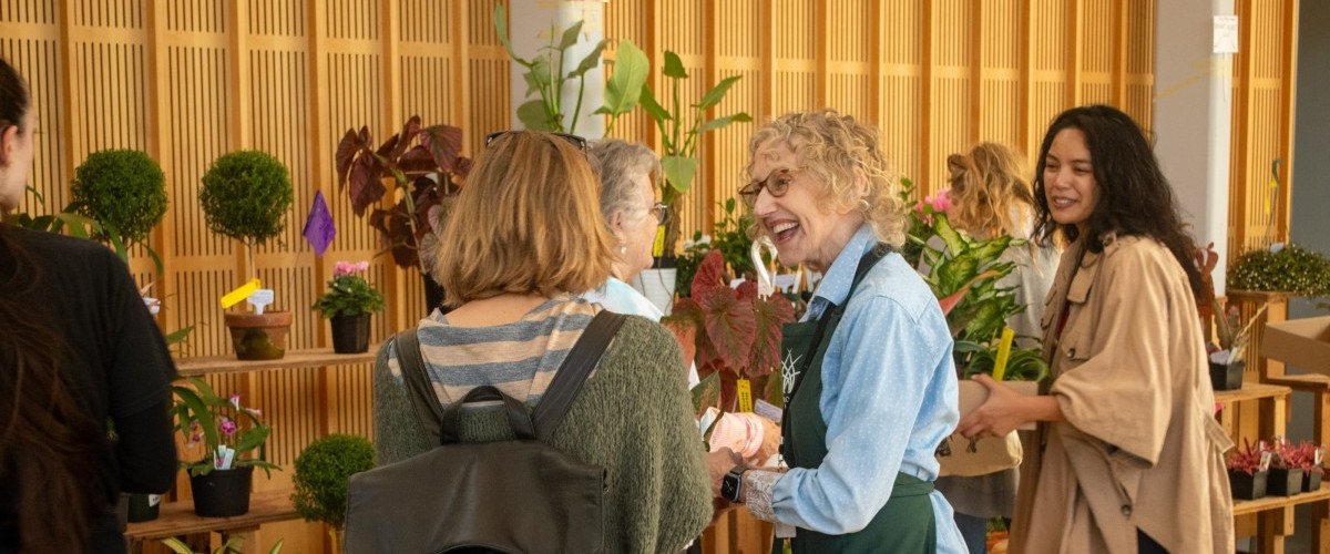 Women looking at plants on sale on shelves and talking with a sales associate wearing a green apron.